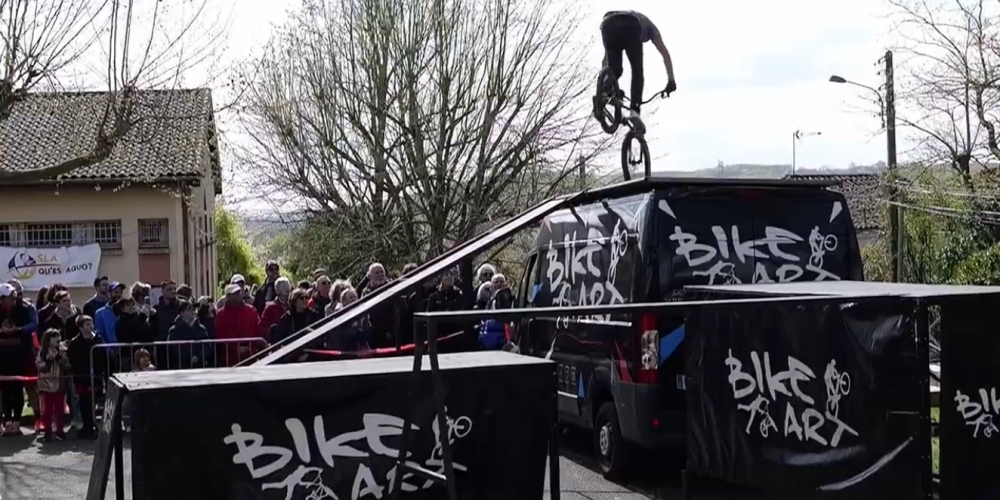 Rider Bike Art en équilibre trial sur le toit du camion, banderole SLA Qu'es Aquò et public de la Cour'SLA Pied 2026
